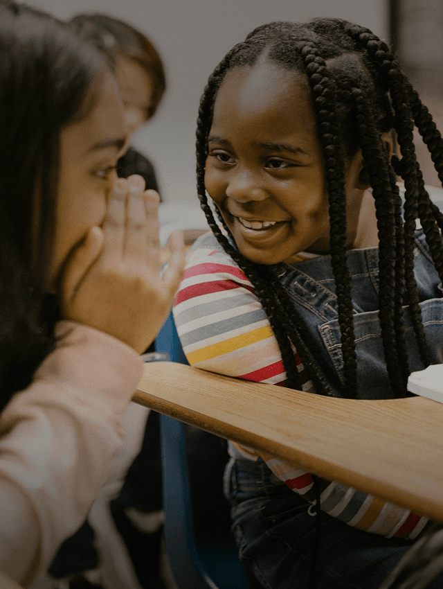 Smiling students whisper to each other as they learn a new language in school.
